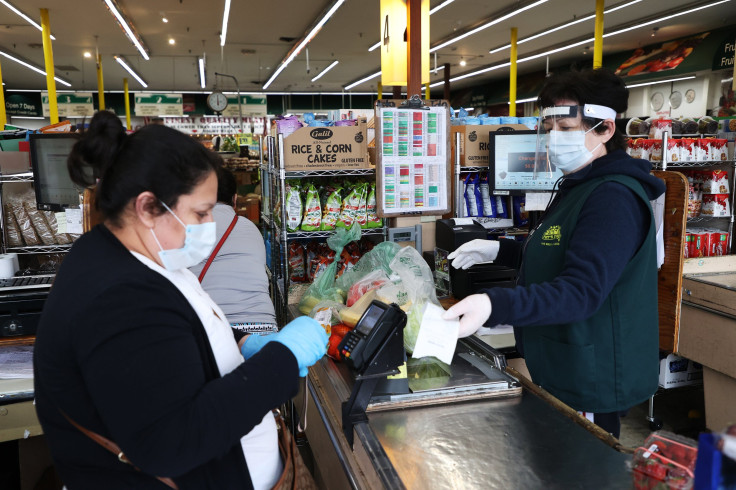  A shopper and cashier both wear masks, gloves and the cashier also has on a plastic visor at the checkout station Pat's Farms grocery store